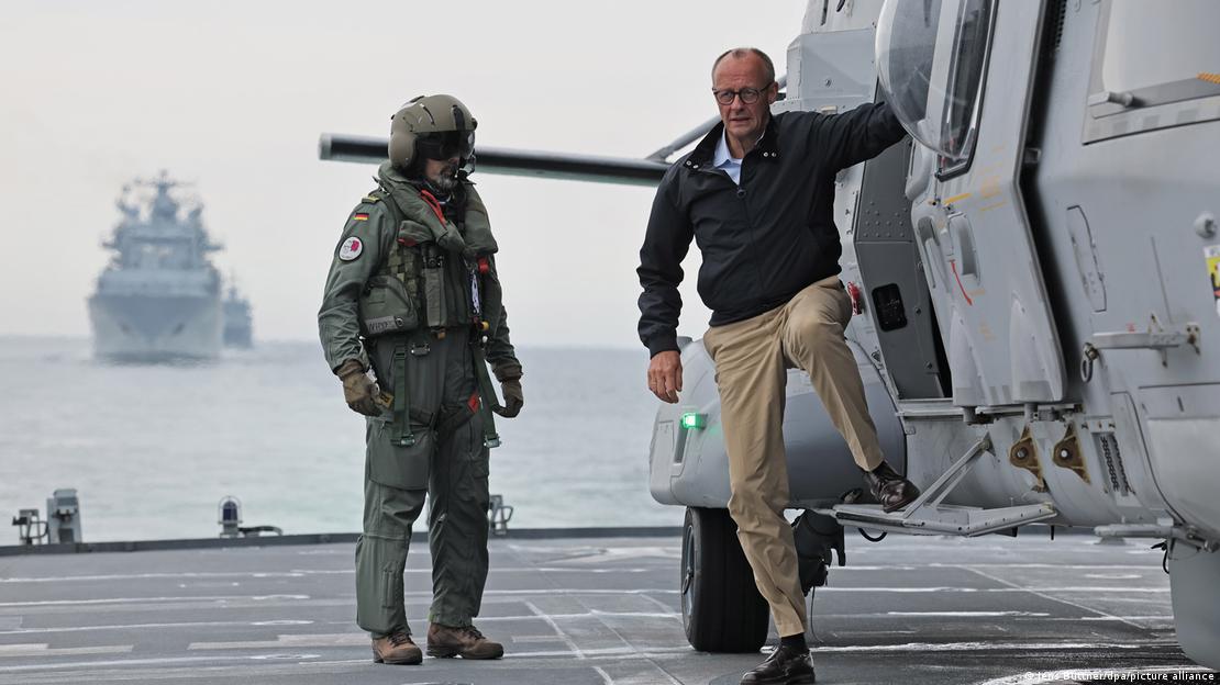 Başbakan Friedrich Merz, helikopterden, Alman Deniz Kuvvetleri'ne ait bir fırkateyne iniyor, yanında kasklı bir asker, arka planda savaş gemileri